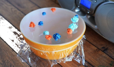 Photo of a bowl covered in clingy plastic wrap with small objects on the film; all set next to a set of speakers