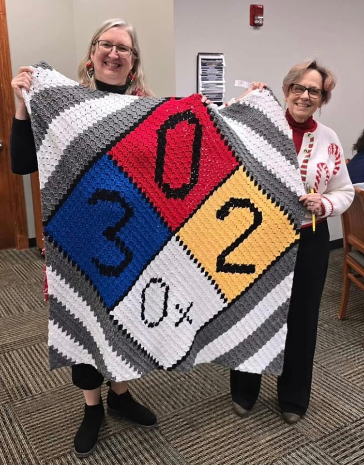 photo of Iota Sigma Pi representative Vivial Sullivan (L) and Janet Feroni (R) holding the lovely crocheted blanket they created. The image on the banket is a familiar diamond-shaped hazard symbol showing the risk levels for concentrated nitric oxide