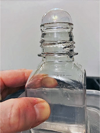 Photo showing a transparent glass bottle with a bubble-like soap film dome over its opening